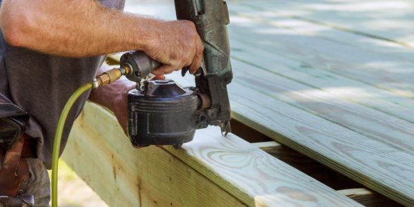 Worker using a nail gun to secure wooden boards during deck construction for a durable and safe structure.