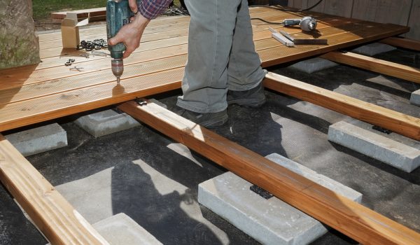 Close-up of a worker using a drill to secure wooden decking boards onto concrete foundation blocks during deck construction.