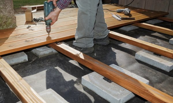 Close-up of a worker using a drill to secure wooden decking boards onto concrete foundation blocks during deck construction.